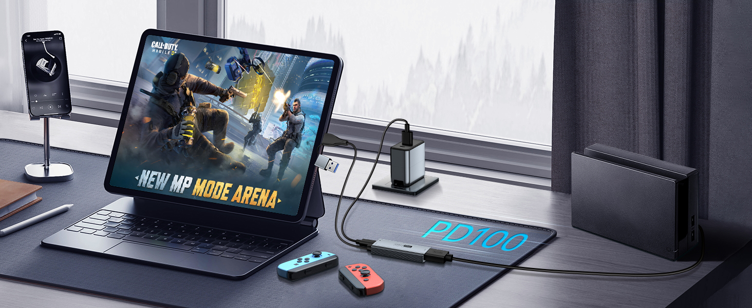 Gaming setup with tablet-like device showing action game, connected to illuminated keyboard. Nearby gaming controller and mouse on desk with window in background.