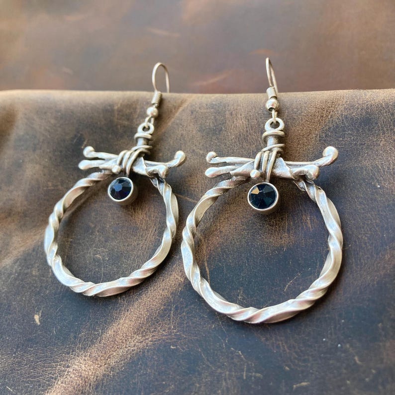 Boho gothic silver-plated twisted hoop earrings with dark blue zircon stones hanging at the center. Rustic wire details accent the top of each hoop. Photo shows the earrings lying flat on a light brown background, taken from a top-down angle.