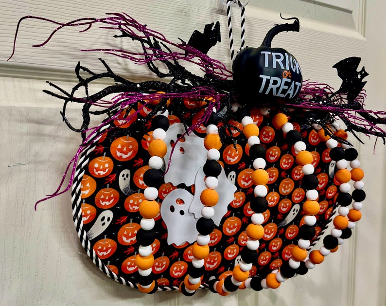 可能包含:一个黑白相间的南瓜形花环,上面镶有橙色和白色的珠子。花环顶部有一个黑色南瓜,上面印有“不给糖就捣蛋”的字样。花环上装饰着黑色蝙蝠和紫色树枝。