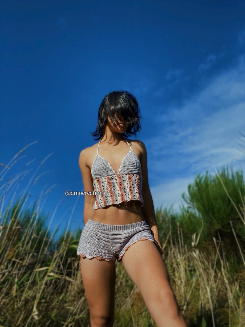 May include: A crocheted two-piece outfit in shades of gray and peach. The top is a halter style with vertical stripes, and the shorts are high-waisted with scalloped edges. The outfit is shown against a backdrop of tall grass under a clear blue sky. The text '@amocreations' is visible on the image.