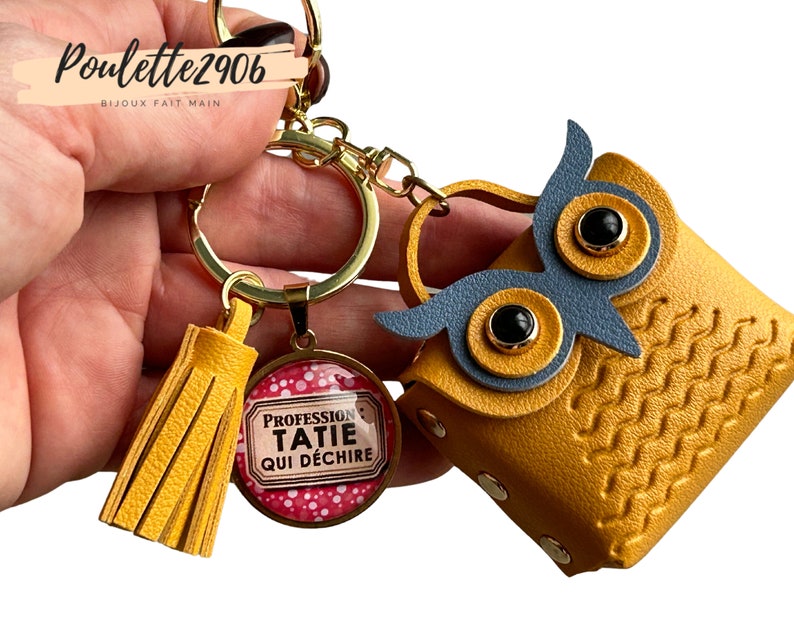 May include: A yellow leather owl-shaped keychain with a gold key ring and a yellow tassel. The owl has black eyes and blue wings. The keychain also has a round glass charm with a pink background and white polka dots. The charm has the text 'Profession Tatie Qui Déchire' in black lettering.