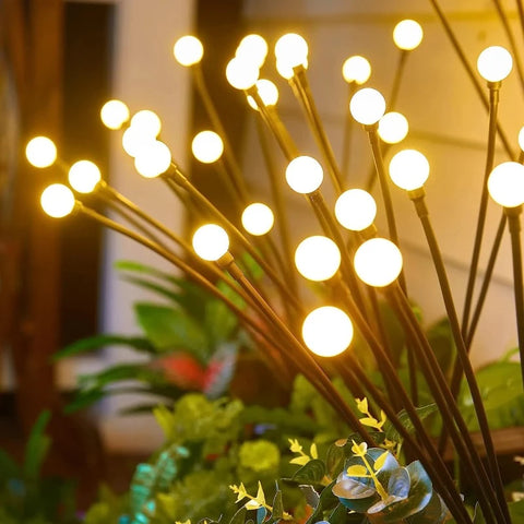 Several slender black stems rise from lush green foliage, each topped with a glowing warm white spherical light. The lights vary in height and create a whimsical, firefly-like effect in a garden setting at dusk or dawn.
