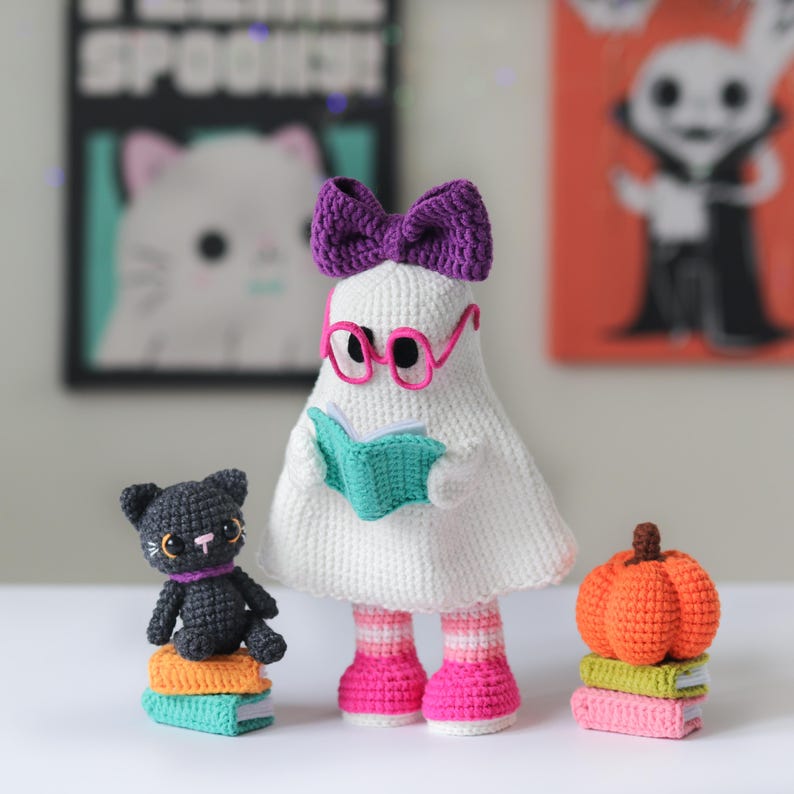 Adorable cute handmade crochet bookish ghost doll with pink glasses, a purple bow, and pink striped boots, holding a teal book – accompanied by a small black crochet cat plush sitting on a stack of books and a bright orange crochet pumpkin. Perfect for Halloween or fall decor. This listing includes a digital download PDF crochet pattern to make your own spooky amigurumi set