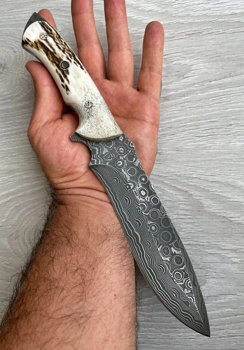 Un cuțit Bowie autentic de Damasc, cu m&acirc;ner din corn de cerb și arțar, forjat &icirc;n 150 de straturi, lucrat manual de un fierar.