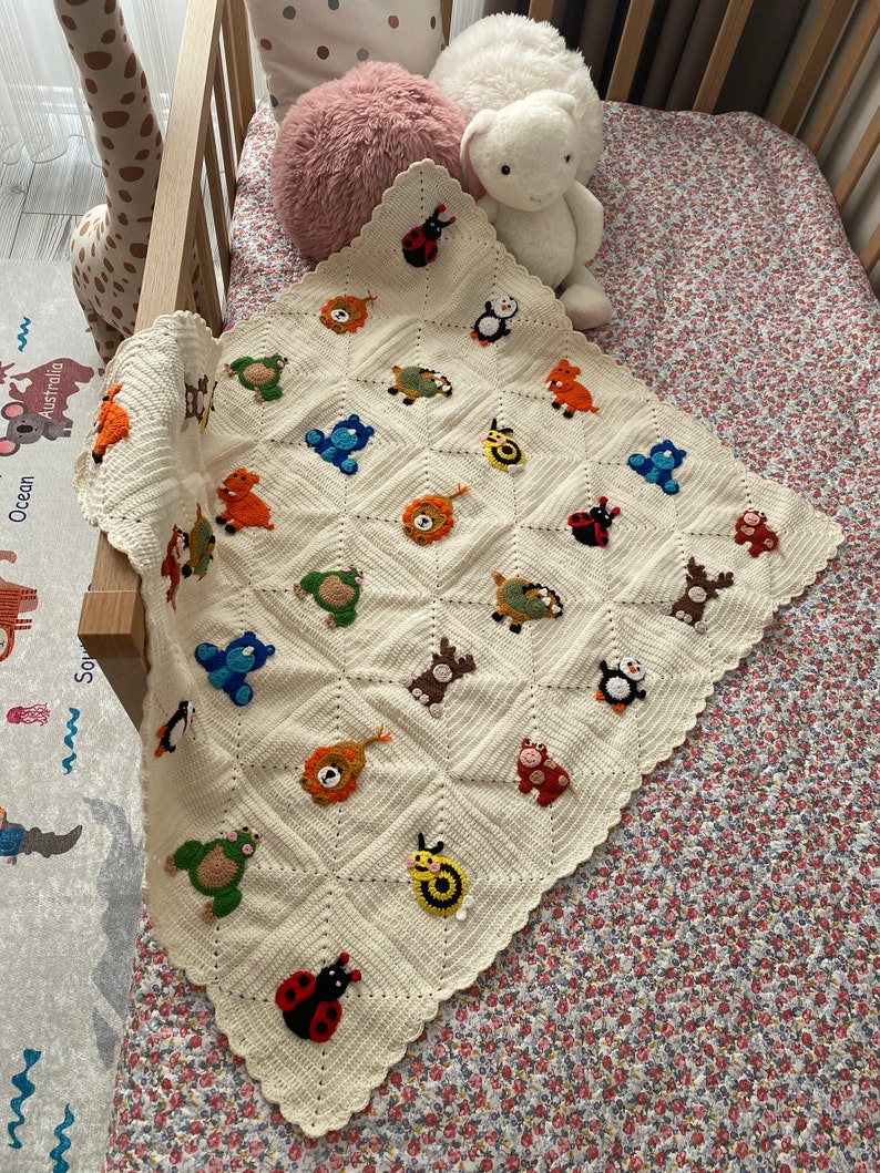 May include: A white crocheted baby blanket with a scalloped edge. The blanket is decorated with colorful animal appliques, including a ladybug, a penguin, a lion, a bear, a turtle, and a snail.