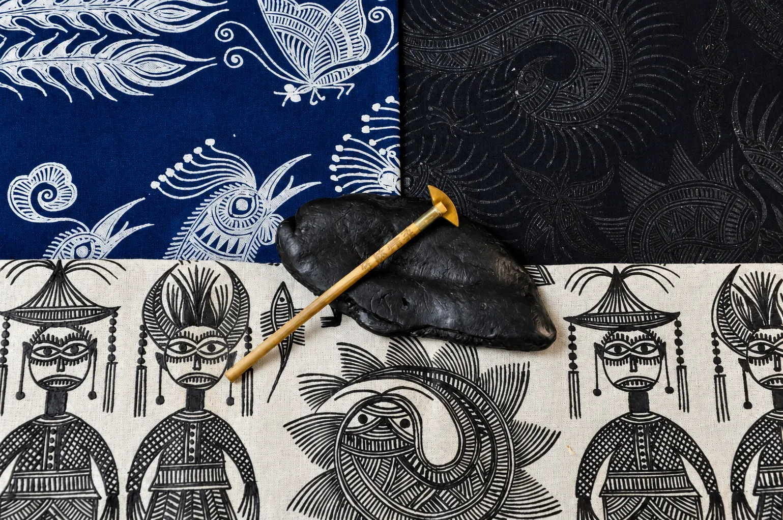 A brass wax tool resting on black wax over indigo and ivory batik textiles with traditional bird and figure motifs