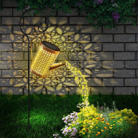 A decorative, golden watering can with intricate cutouts hangs from a black shepherd's hook in a garden setting. Solar-powered lights within the watering can illuminate, casting floral patterns onto a brick wall behind. A stream of small, warm-toned lights appears to pour from the watering can's spout onto a patch of colorful flowers and green foliage below. The scene suggests a whimsical and illuminated garden decoration at dusk or dawn.