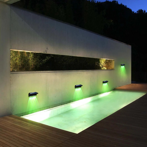 Four solar-powered fence lights are mounted along a low wall, illuminating a wooden walkway at dusk or night. The lights cast beams downwards onto the path.