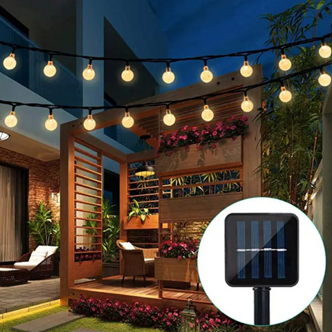Two strings of round, warm-toned lights illuminate a patio area with wooden structures, plants, and outdoor furniture at dusk. A solar panel for the lights is shown in a circular inset.
