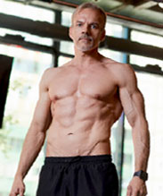 A muscular man with gray hair stands shirtless in a gym.