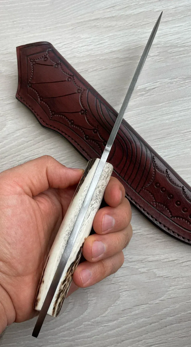 Un cuțit Bowie autentic de Damasc, cu m&acirc;ner din corn de cerb și arțar, forjat &icirc;n 150 de straturi, lucrat manual de un fierar.
