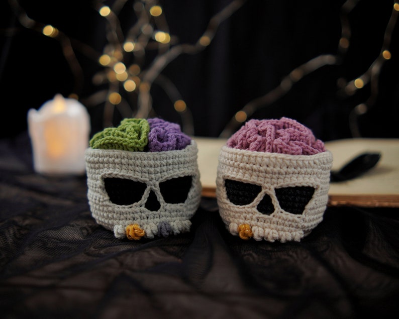 May include: Two crocheted skull shaped containers with a brain-shaped crocheted insert. One container has a green and purple brain insert and the other has a purple brain insert. The containers are on a black and white textured surface.