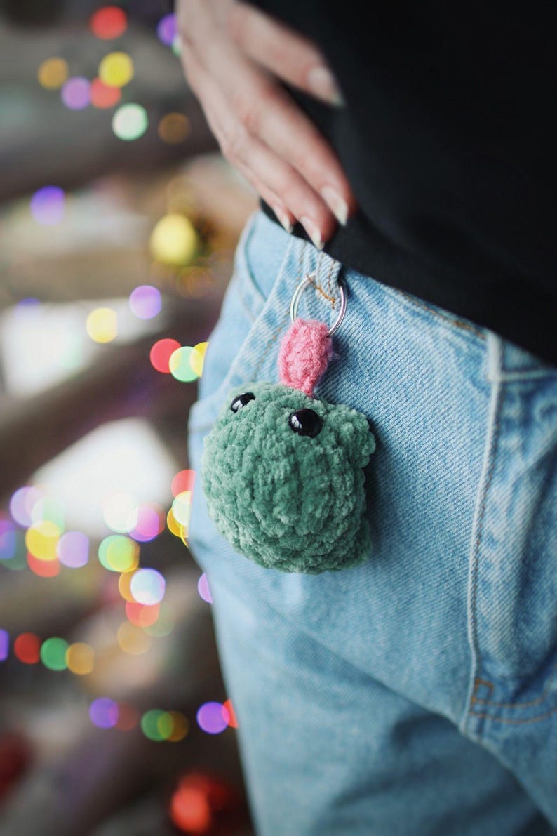 May include: A green crocheted keychain with pink yarn accents and black button eyes.