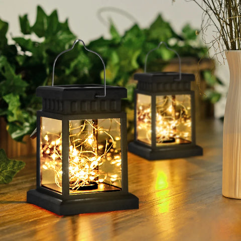 Two solar-powered fairy lanterns with black ABS frames and crackle-style LED string lights inside, casting a warm glow on a wooden surface. The lanterns feature a hanging handle, making them suitable for outdoor or indoor decorative use. Green plants and a vase are visible in the background, creating a cozy ambiance.