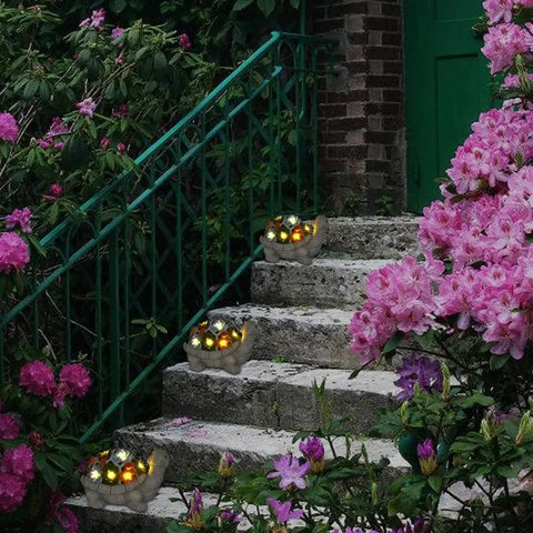 Three decorative gray turtle statues with segmented shells, each containing small glowing lights, are placed on stone steps leading up to a green door. Pink and purple flowers and green foliage surround the steps.