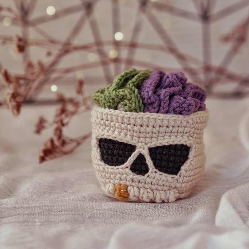 May include: A crocheted skull-shaped basket with a purple and green crocheted flower inside. The basket is beige with black eye details and a yellow button on the bottom.