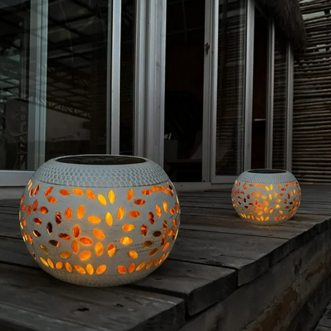 Two round, white outdoor lanterns with decorative cutouts emitting a warm orange glow sit on a wooden surface at dusk or night. The lanterns have dark, flat tops, likely solar panels. A building with large windows is visible in the background.