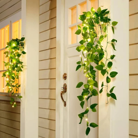 Artificial green vines with integrated warm white fairy lights hang on a white front door and near a window of a house with beige siding at dusk or night.