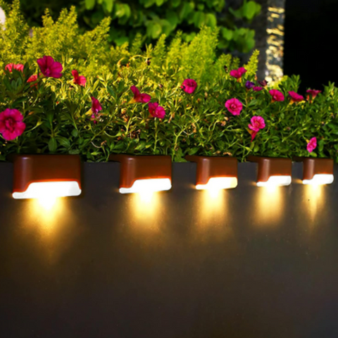 Five small, brown, semi-circular solar lights are mounted in a row along the base of a dark wall at night, emitting a warm white upward glow. Planter boxes filled with green foliage and vibrant magenta flowers sit above the lights. The scene shows illuminated garden wall lights enhancing the plants.