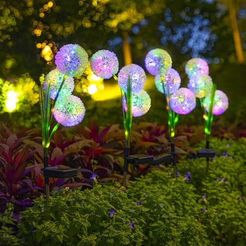 Several solar-powered garden lights that resemble stylized dandelions. They are placed among flowers and greenery in a front yard at dusk or dawn, casting a soft, multicolored glow. The house's facade is visible in the background.