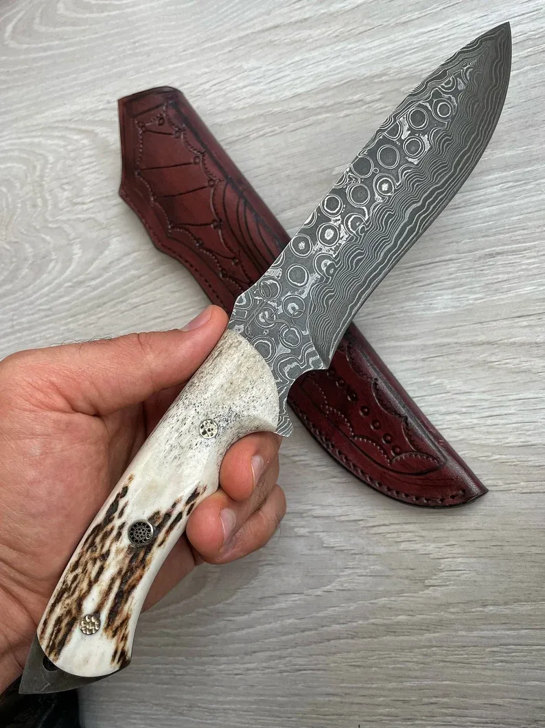 Un cuțit Bowie autentic de Damasc, cu m&acirc;ner din corn de cerb și arțar, forjat &icirc;n 150 de straturi, lucrat manual de un fierar.