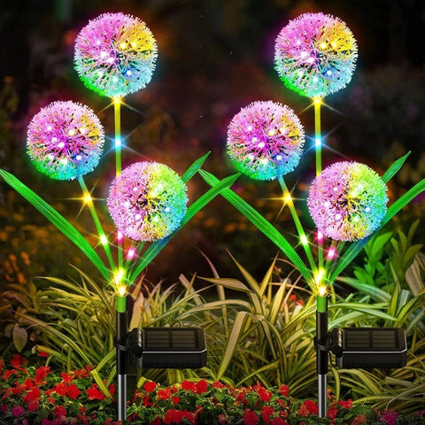 Six solar-powered garden lights are arranged in two rows. Each light features a spherical, dandelion-like head emitting a vibrant, multicolored glow, including pink, yellow, blue, and green. They have green, leaf-shaped accents and black solar panels at the base. The lights are placed among low-lying greenery and small red flowers.