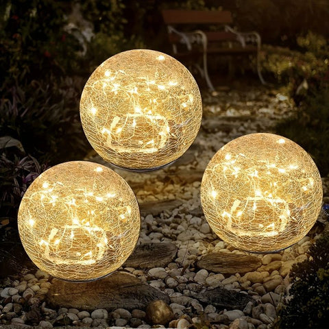 Three solar-powered crackle glass globe lights emit a warm white glow in an outdoor garden setting at night. The lights are placed on a pebble pathway, creating a cozy and decorative ambiance. A wooden bench and lush greenery are visible in the softly illuminated background.