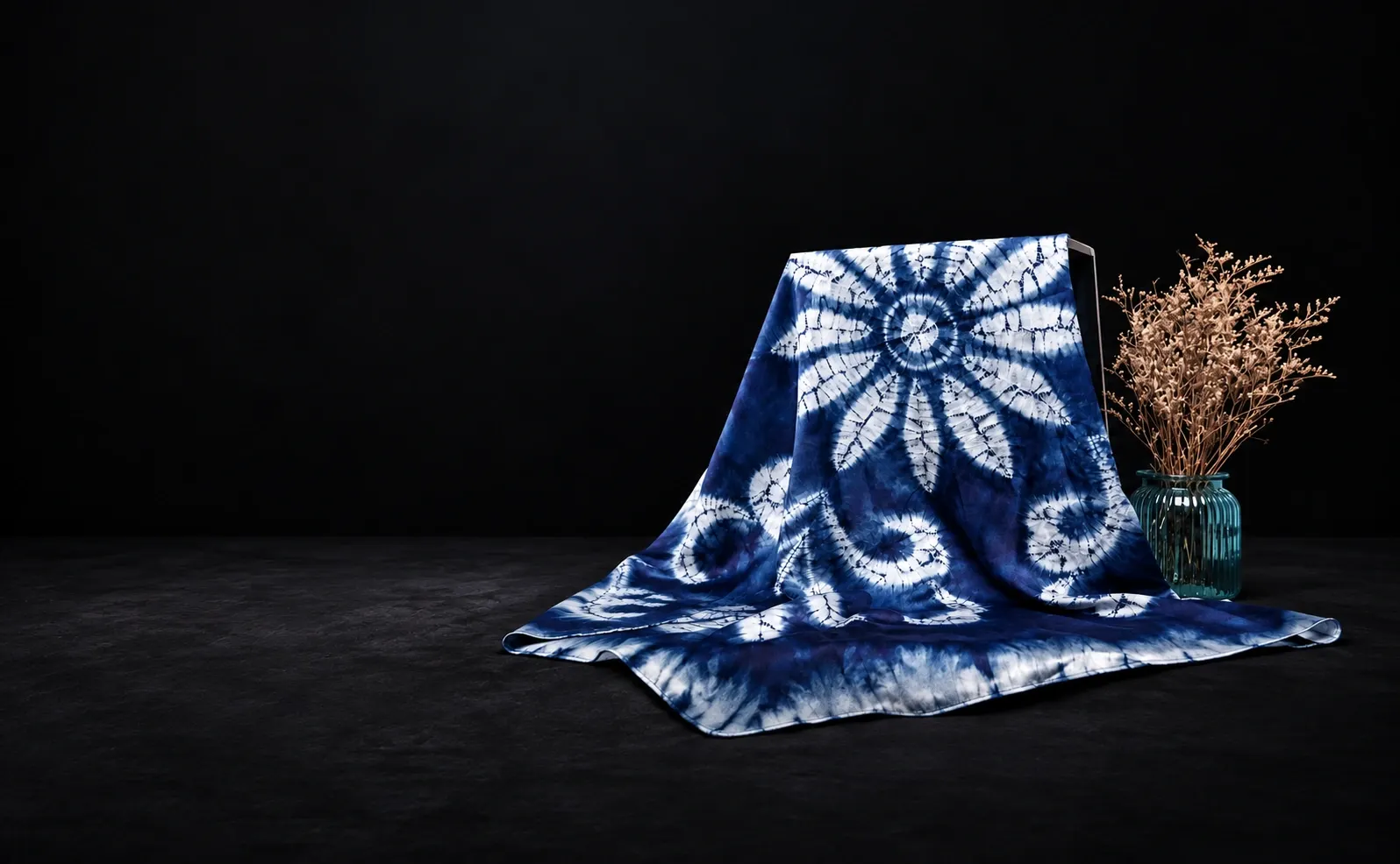 Blue-and-white radial tie-dyed cloth beside a green glass vase of dried flowers