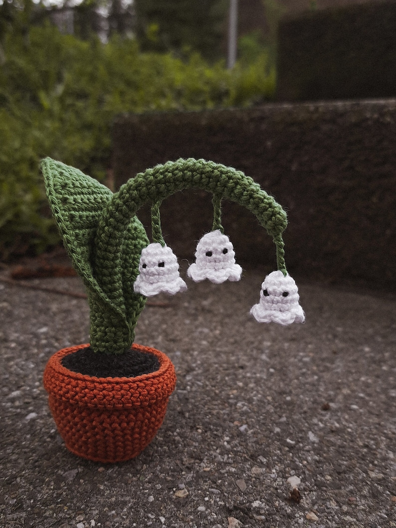 May include: A crocheted lily of the valley plant in a terracotta pot. The plant has white flowers with black eyes and green leaves.
