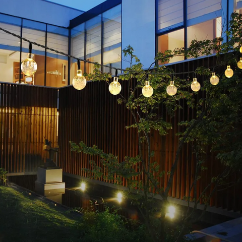 A string of round, glowing lights hangs across an outdoor space with a wooden fence and greenery. A dark pond reflects the lights. Modern buildings with large windows are visible in the background.