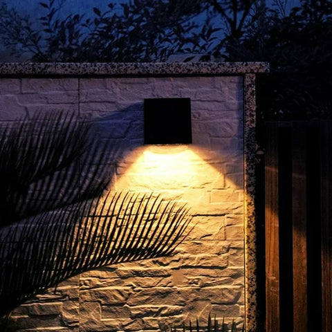 A black solar-powered square wall lamp is mounted on a white brick wall, casting a warm yellow light in a triangular pattern. A palm leaf's shadow is visible on the wall, adding depth to the nighttime outdoor setting.