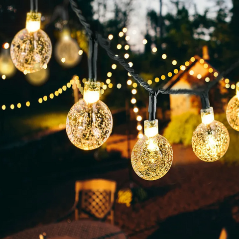 A close-up shows a string of round, textured lights glowing with a warm light in an outdoor setting. Other strings of smaller lights are blurred in the background. A glimpse of a patio chair is visible below.