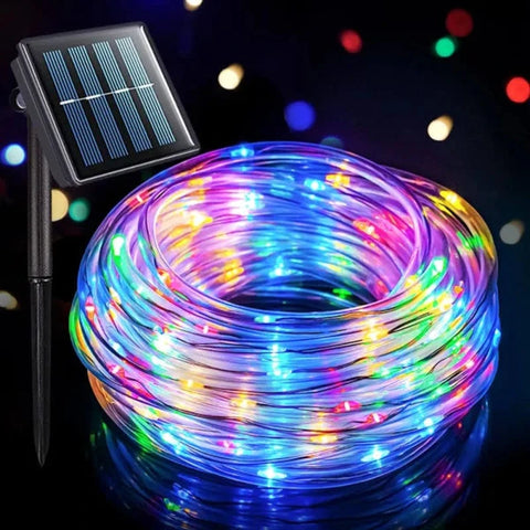 A coiled length of flexible rope light with multiple colored LED bulbs is shown with its attached solar panel and ground stake against a dark background with colorful bokeh lights. The rope light displays a mix of blue, pink, yellow, and white lights.