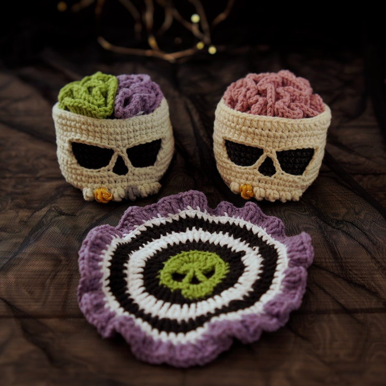 May include: Three crocheted items: two skull-shaped baskets with green and pink brains inside, and a purple and black circular coaster with a green skull in the center.