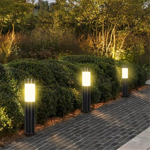 Three cylindrical outdoor pathway lights illuminate a brick walkway bordering lush green bushes at dusk or dawn. The lights have dark bases and frosted white diffusers, emitting a warm, upward glow. The pathway curves gently into a garden area with trees in the background.