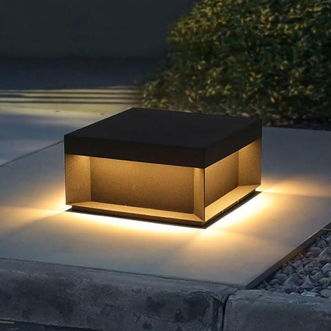 A modern, square outdoor pillar light with a dark top and glowing warm-toned panels, illuminating a concrete surface at night.