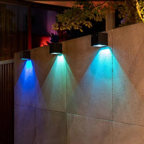 Three black solar-powered square wall lamps are mounted on a concrete wall, emitting blue and cyan lights in a triangular pattern. The outdoor setting is illuminated by ambient lighting, with trees and a building visible in the background.