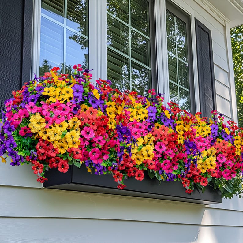 May include: A vibrant window box overflowing with colorful artificial flowers. The arrangement features a mix of red, pink, yellow, orange, and purple blooms, creating a cheerful display. The flowers are set in a black rectangular planter, adding a contrast to the bright colors.