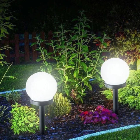 Two spherical white solar garden lights on black stakes illuminate a mulched garden bed with various plants at night.