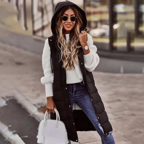 Eine stilvolle Frau in einer langen schwarzen Pufferweste, kombiniert mit einem weiÃŸen Pullover und Jeans, sowie Accessoires wie einer Sonnenbrille und einer Handtasche.