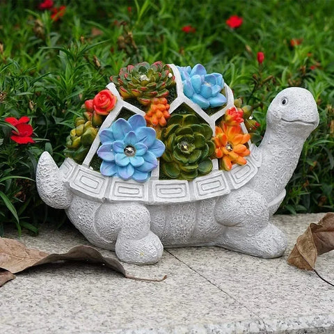 A gray decorative turtle statue with a segmented shell holding colorful artificial succulents is displayed outdoors on a stone surface with fallen leaves. Green foliage with red flowers is visible in the background.