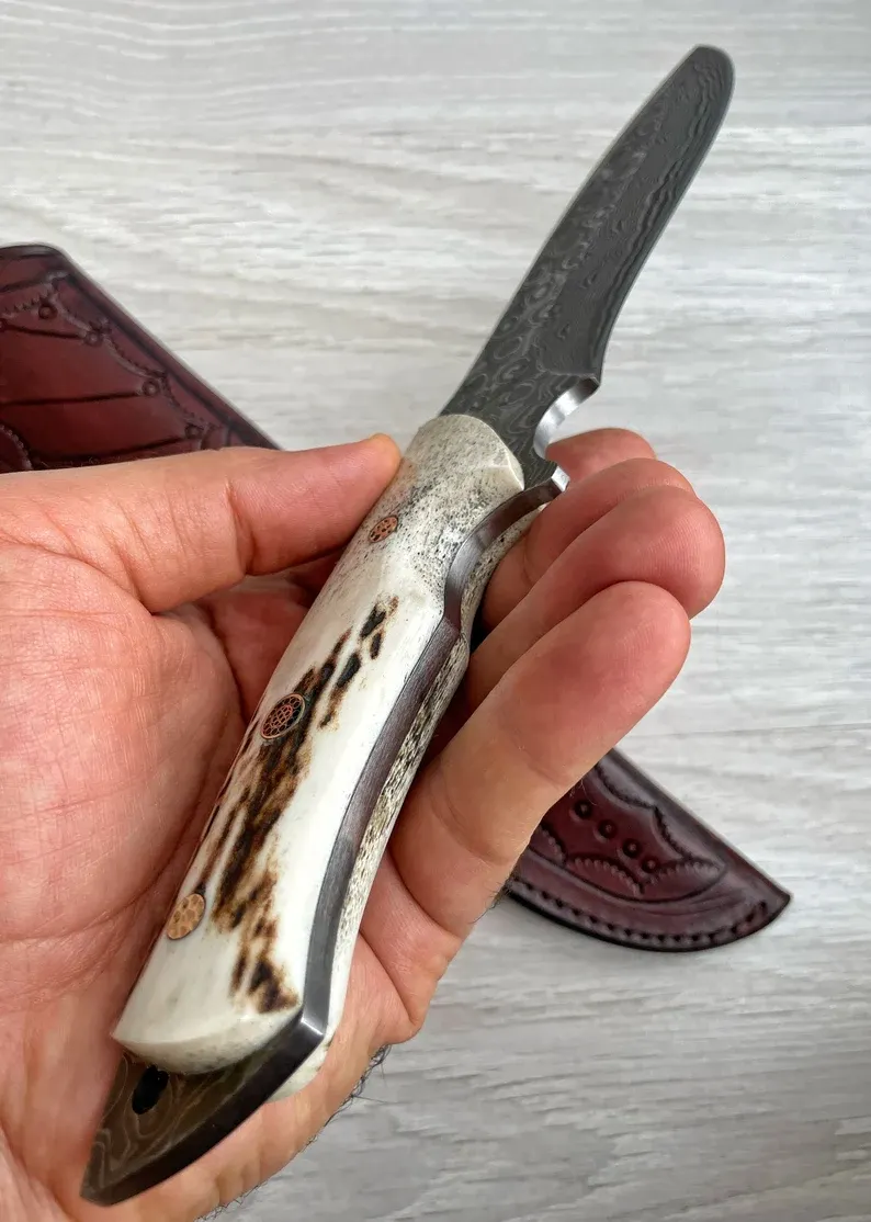 Un cuțit Bowie autentic de Damasc, cu m&acirc;ner din corn de cerb și arțar, forjat &icirc;n 150 de straturi, lucrat manual de un fierar.