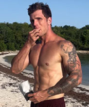 Shirtless man with tattoos standing on a beach and eating.