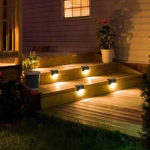 Four small, dark, rectangular solar lights are mounted along the sides of wooden outdoor steps at dusk or dawn, emitting a warm white downward glow. The steps lead up to a house with a lit window and potted plants. Grass is visible at the bottom of the steps. The scene shows illuminated outdoor stairs.