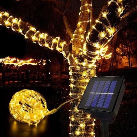 A string of warm white fairy lights is wrapped around the trunk and branches of a tree at night. The lights are also coiled in a cluster on the ground near a small solar panel on a stake, which likely powers them. The background is dark with blurred lights suggesting an outdoor setting.