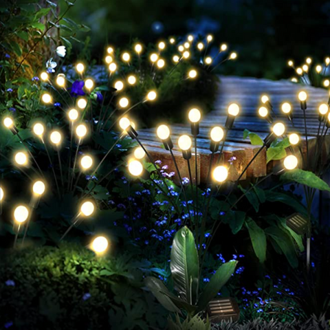 Several slender black stems rise from lush green foliage, each topped with a glowing warm white spherical light. The lights vary in height and create a whimsical, firefly-like effect in a garden setting at dusk or dawn.