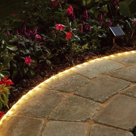 A flexible rope light emitting a warm white glow outlines a curved stone pathway at dusk or dawn. The rope light sits along the edge of the path, next to dark mulch and vibrant pink flowers with green foliage. A small solar panel on a stake is visible near the flowers, likely powering the rope light.