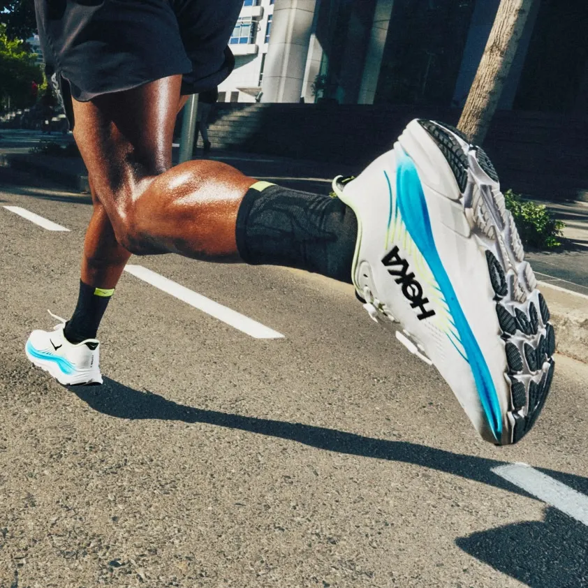 Runner's legs in mid-stride. White HOKA running shoes, one foot airborne on road.