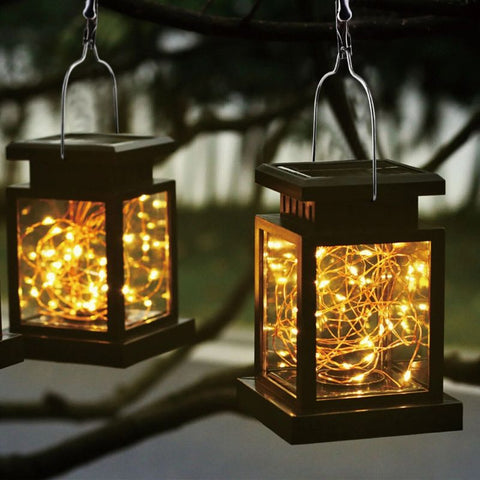 Two solar fairy lanterns with black ABS frames and delicate LED string lights inside, hanging from tree branches at dusk. The lanterns emit a warm golden glow, creating a magical and inviting outdoor ambiance. The solar panels on top ensure energy-efficient lighting.
