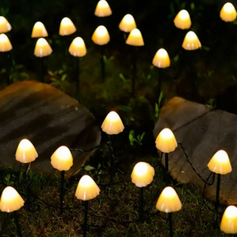Numerous small, mushroom-shaped solar lights with translucent caps emit a warm white glow in a garden at night. The lights are on thin black stakes and are scattered among rocks and dark foliage.